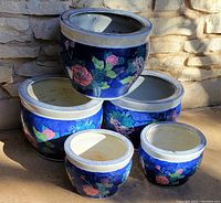 Five round ceramic flower pots with bright blue background and colorful flowers, stacked outdoors in sunlight against stone wall. Pots have solid bottoms, no drainage holes, and white rims with decorative pattern.