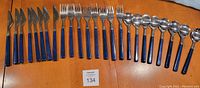 Flatware set spread in a single row on wooden table showing knives, forks, teaspoons and tablespoons all with blue plastic handles.