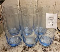 Six tall cylindrical blue tinted glasses standing grouped on a speckled countertop, each showing a blue tint predominantly at the base.