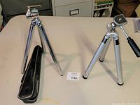 Two camera tripods on table, chrome tripod with black soft case on left, silver tripod on right with handle.