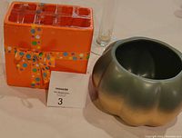 Orange square ceramic flower vase with polka dot bow design and green pumpkin-shaped ceramic planter side by side on table with maxsold number card.