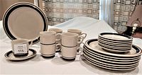 Full set displayed including stacked plates, cups stacked and arranged with saucers. Beige stoneware with brown bands.