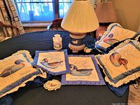 Duck themed throw pillows with ruffled edges featuring detailed embroidered duck images, a lamp with wooden base decorated with a white duck figure and a cream-colored pleated lampshade on a table beneath window with curtains.