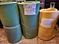 Two green twin-size egg crate foam mattress toppers rolled and stacked, secured with rubber bands next to a yellow/beige full-size egg crate foam mattress topper also rolled and secured.