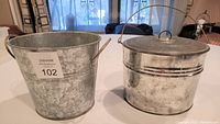 Two galvanized round metal containers on a table, one with riveted handles and one with a lid and wire handle