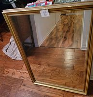 Full view of gold framed rectangular mirror with reflective surface, placed on wooden floor leaning against wall.