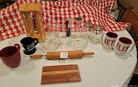 Photo showing assortment of kitchen items including mugs, measuring cup, citrus juicers, rolling pin, cutting board, spice rack, mason jars placed on red and white checkered tablecloth.