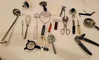 Various kitchen utensils including ladles, whisks, potato masher, can openers, ladle, peelers, sifter, and others laid out on white cloth.