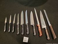 Photo of ten kitchen knives arranged in a row with varying handle styles; six with black handles marked Chefstyle, others marked Farberware, MightyOak, Victorinox and one unknown. Knives show signs of use and dull blades.