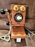 Front and side view of wooden rotary phone replica on brown couch showing rotary dial, handset, and brass bells.