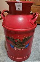 Front view of red antique milk can displaying American eagle decal and handles.