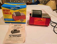 Chicago rotary tumbler next to original box and instruction booklet on white cloth.