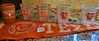 Photo showing four clear plastic glasses each with a red Texas Longhorn logo, one brown insulated Texas Longhorn cup, and an orange Texas cloth with logo.