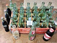 Photo showing the original red Coca-Cola wooden crate holding 24 various Coca-Cola glass bottles, some empty, some sealed. Also visible two green Dr. Pepper bottles and a red Coca-Cola wall-mount metal bottle opener.