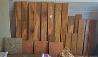 Multiple wood flooring boards with natural grain patterns, Laminate and engineered wood, stacked vertically against wall, alongside three Saltillo floor tiles and one decorative stone-pattern tile on the floor.