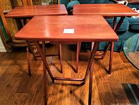 Three wooden folding TV trays shown standing unfolded on a hardwood floor, showing their reddish-brown flat tops and foldable legs.