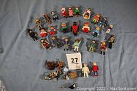 Photo showing 26 Playmobil action figures arranged in rows on a blue fabric surface, including knights, soldiers, and civilian characters, with several accessories like helmets, weapons, and shields in front.