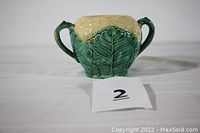 Front view of the green and creamy yellow Etruscan Majolica cauliflower shaped sugar bowl with two handles, sitting on a white textured cloth.