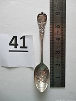 Photo of the full sterling silver souvenir spoon placed next to a ruler to show length of approximately 6 inches and number card '41'. Spoon features figure engravings on bowl and an ornate handle.