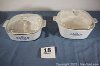 Both casseroles side by side on blue surface, each with clear glass lid and Blue Cornflower design on white ceramic body.