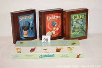 Four board games: Stratego, Yahtzee, Clue in wooden bookshelf boxes standing upright behind Cocktailopoly box lying flat on table.