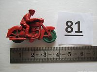 Left-side view of red rubber toy police motorcycle with green wheels beside ruler for size reference.