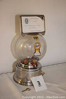 Front view of the gumball machine showing metal base, plastic globe filled with multicolored gumballs, Ford logo, 10¢ sticker, Kiwanis label at top and included key on table.