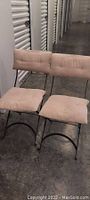 Pair of wrought iron balcony chairs with light beige padded cushions on seat and backrest placed side by side on concrete floor.