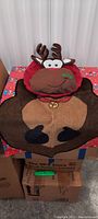 Brown and red fabric toilet seat cover shaped like a reindeer on top of matching floor mat.