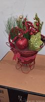 Photo of red and green glittered Christmas decoration in a red wire container showing pine needles, ornaments, and beads.