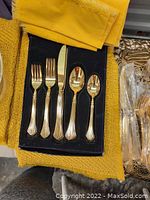Set of 5 gold-toned silverware pieces (forks, knife, spoons) displayed in black box on yellow cloth.
