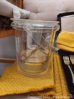 Photo of a clear plastic ice bucket with gold trim on a yellow textured mat, with a clear handle lifted up.
