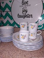 Photo showing a set of white ceramic dinnerware including plates, bowls, and cups with fruit pattern on cups and blue zigzag design on plates and bowls.
