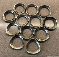 Top-down view of all 12 silver plated napkin rings arranged on a flat surface showing their irregular, rounded rectangular and oval biomorphic shapes with shiny silver surface and slight tarnishing.