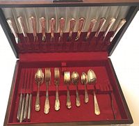 Photo showing the organized cutlery set in a wooden case with red felt lining, displaying knives, forks, spoons, a ladle, and a butter knife.