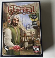 Front cover of the Istanbul board game box showing the game title, publisher logos, and artwork of a merchant holding rubies in a market setting.