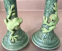 Close-up of the raised frog detail on one green pottery candlestick showing textured bark design and yellow-green frog figure.