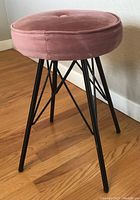 Photo of an Eames style Eiffel base stool showing a round pink fabric cushioned seat and black metal legs with cross-braced construction.