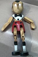 Full front view of the Pinocchio wooden figurine showing painted facial features, black hair, white shirt with black bow tie, red shorts, and articulated limbs.