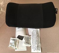 Black lumbar support cushion with ergonomic design, fabric material, small size, and rectangular shape. Includes control switch for massager with settings OFF, LO, and HI. Visible original paperwork and packaging inserts are included in the background.