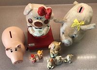Photo showing 4 piggy banks: large pink ceramic, vintage pig with red bow, plush piggy bank with yellow bow, and plain pink piggy bank. Also shows 5 small pig figurines in front.