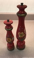Two red wooden grinders with hand painted floral motifs, one taller and one shorter, shown standing upright on a beige countertop.