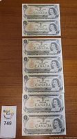 Photo showing all seven 1973 Bank of Canada one dollar bills laid out flat in two columns on a wooden surface.