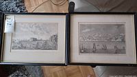 Two framed art prints placed side by side on the floor, showing city views with ships and buildings, each framed in black with matte borders.