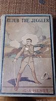 Front cover of the book titled "Rujub the Juggler" showing an illustrated man in military attire with a rifle, with visible wear and stains on cover.