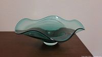 Angled side view showing the flowing wavy organic form of the teal-green glass bowl on a wooden surface.