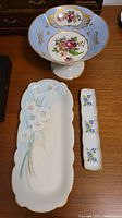 Photo of three Limoges porcelain dishes on wooden surface: footed bowl, rectangular dish, narrow dish with floral decorations and gold trim