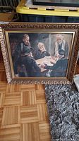 Full view of the framed painting depicting five elderly men gathered around a table, placed on a wooden floor near a rug.