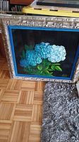 Framed square painting with silver ornate frame leaning against a yellow box, depicting blue hydrangea flowers on black background.