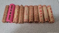 Ten rolls of pennies wrapped in red and brown paper with denomination marked as 50 cents each, lying side by side on a speckled countertop. A pink device (coin counter) is placed next to the rolls.